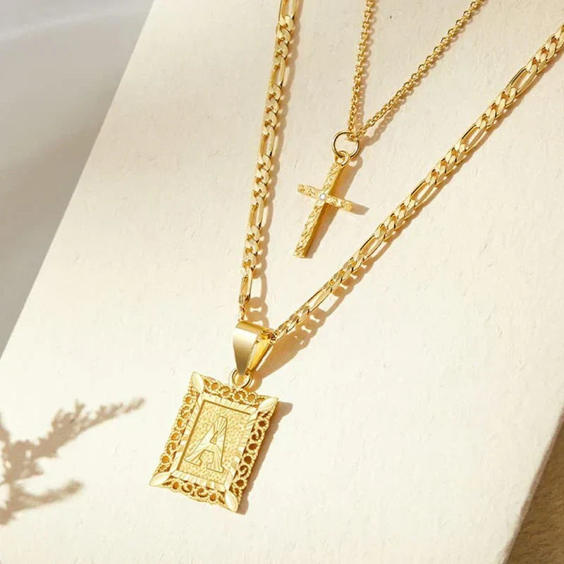 Layered gold cross and square letter pendant necklaces resting on a cream surface in soft shadow 2