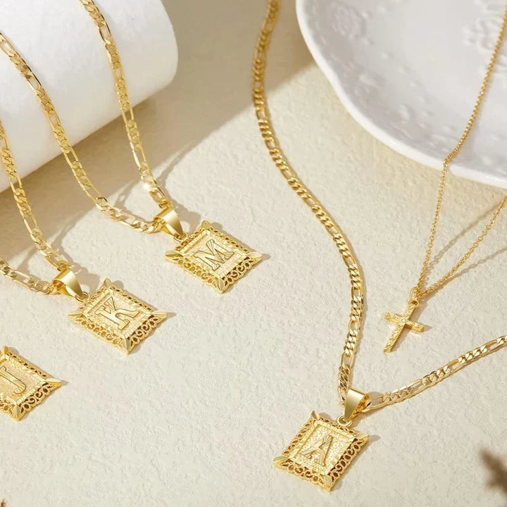 Several gold square letter pendant necklaces and a cross necklace arranged on a cream surface beside a white dish 3