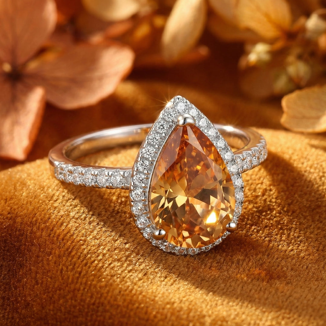 Silver-tone halo ring with amber pear-shaped stone resting at an angle on mustard velvet beside dried flowers 1