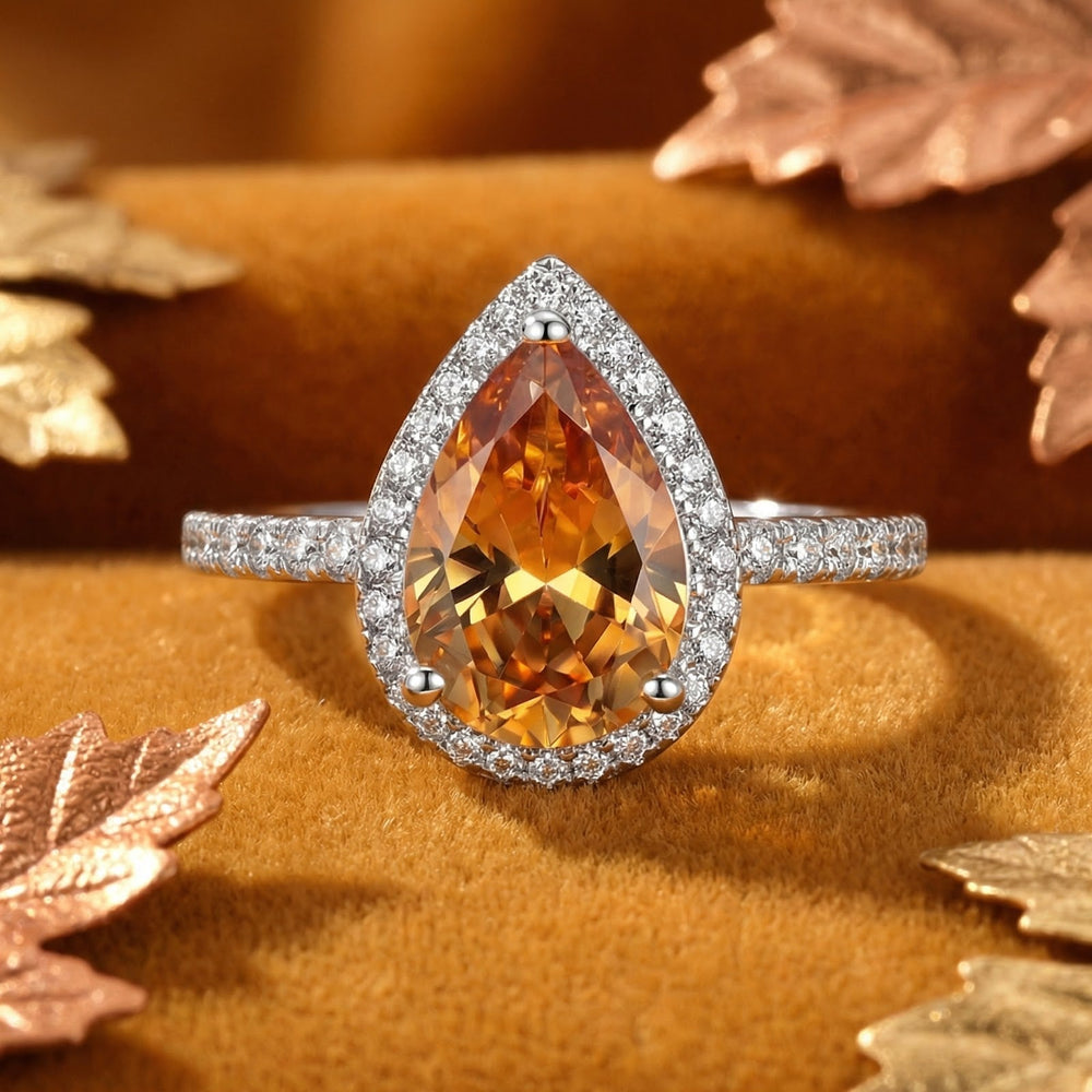 Front view of the silver-tone ring with an amber pear-shaped center stone, sparkling halo and band, among metallic leaves 2