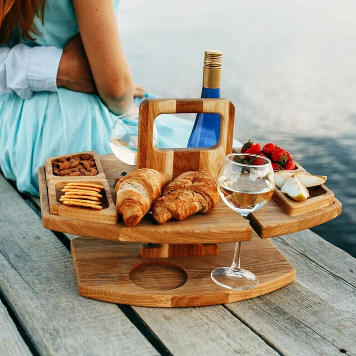 Wooden table on a lakeside dock holding croissants, almonds, crackers, strawberries, pear slices, a wine glass, and a blue 4