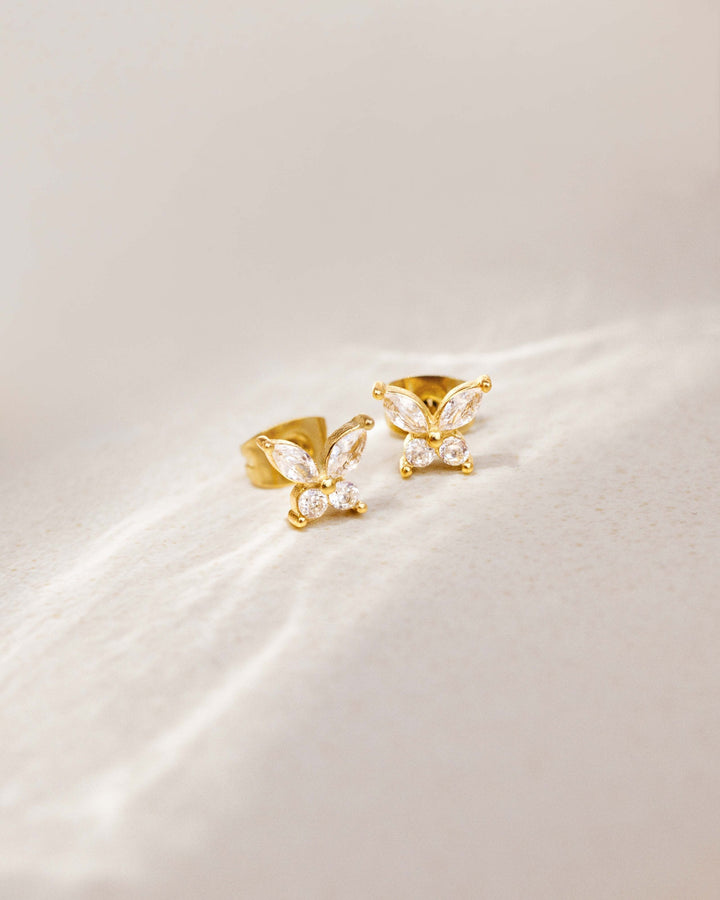 Gold-tone butterfly studs with clear stones resting on a soft cream surface with light streaks 10