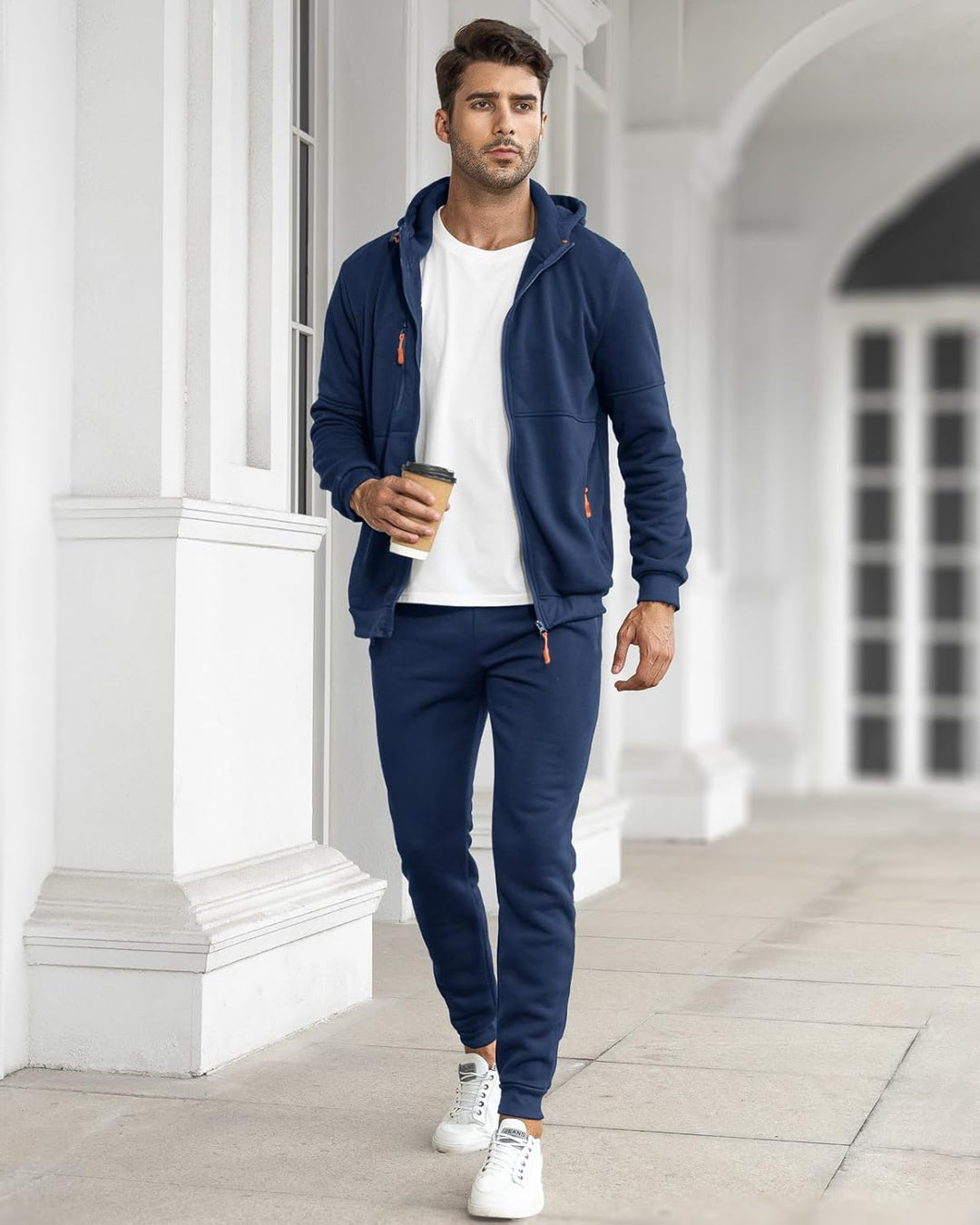 Man in a navy tracksuit with the jacket open over a white tee, holding a coffee 4