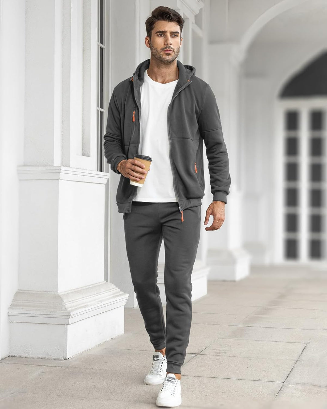 Model in the open gray hoodie over a white tee, holding a coffee cup along a white arcade 13