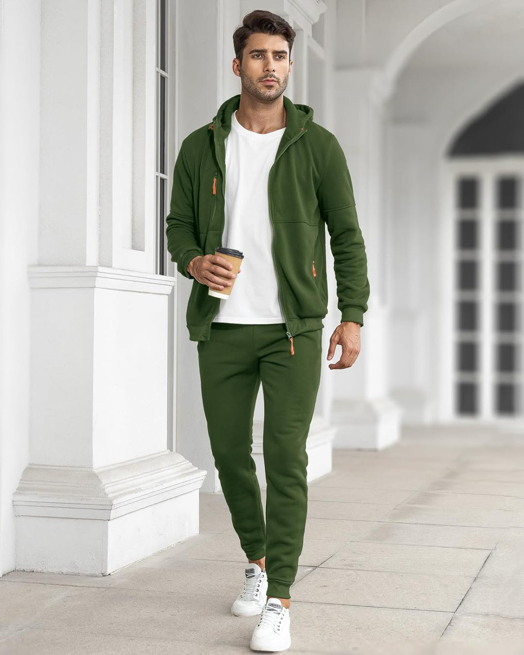 Model in the open green hoodie over a white tee, holding a coffee cup beside white columns 19