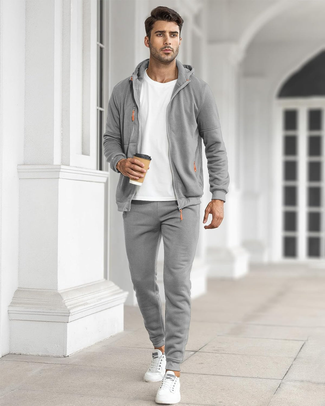 Man in the light gray tracksuit, jacket open over a white tee, walking past white columns with coffee 16