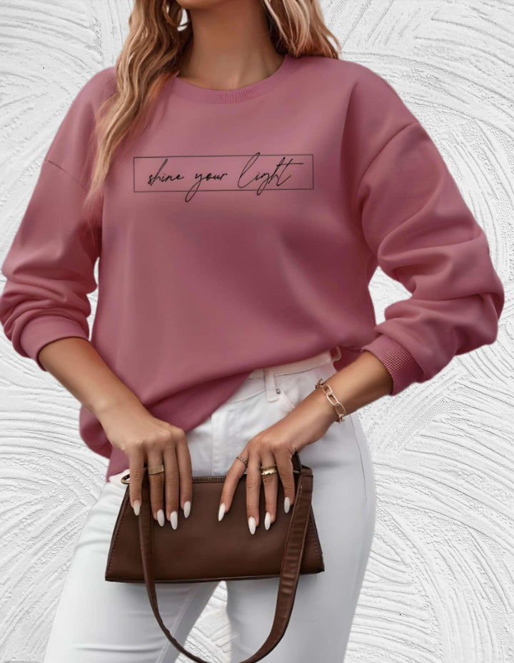 Frau in rosa Pullover mit "shine your light"-Aufdruck, weiße Hose, braune Handtasche.