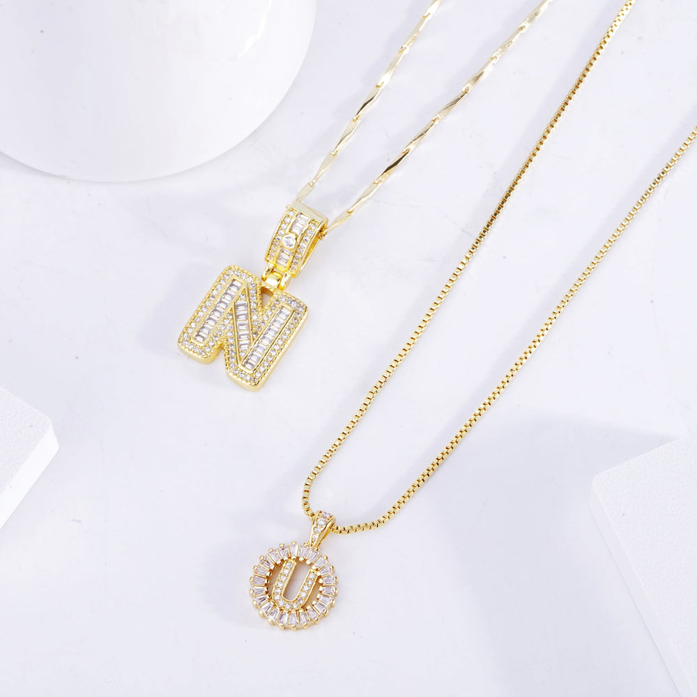Gold double necklace laid flat on a white surface, showing crystal-set block and round initial pendants up close 2
