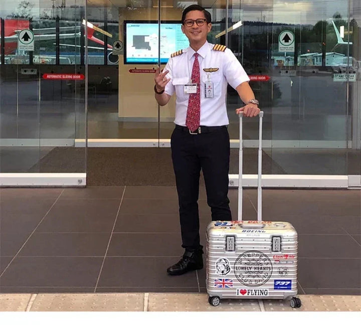 Airline pilot in uniform smiling beside a silver ribbed case covered in travel stickers 4