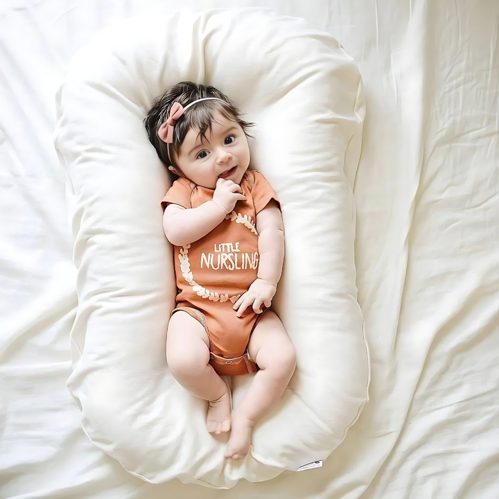 Baby with a pink hair bow and orange bodysuit lies on a cream lounger on bedding 5