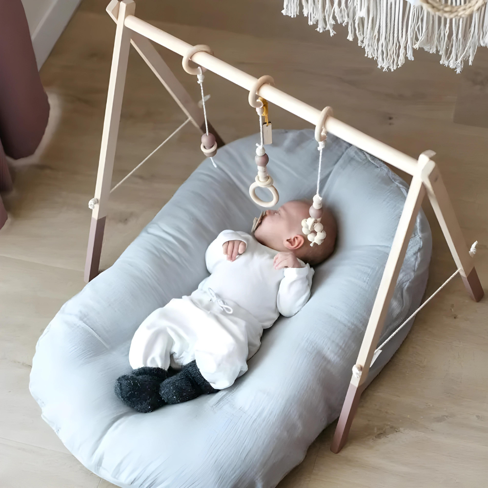 Baby in white outfit lies on a pale blue lounger beneath a wooden play gym 6