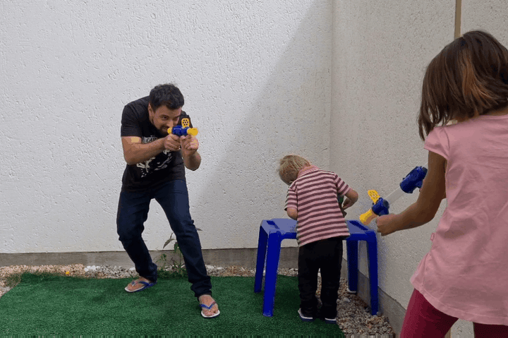 Man and two children play outdoors with blasters beside a white wall and a blue stool 9