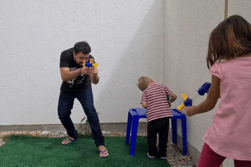 Man and a girl aim blue and yellow blasters outdoors while a toddler stands at a blue stool 10