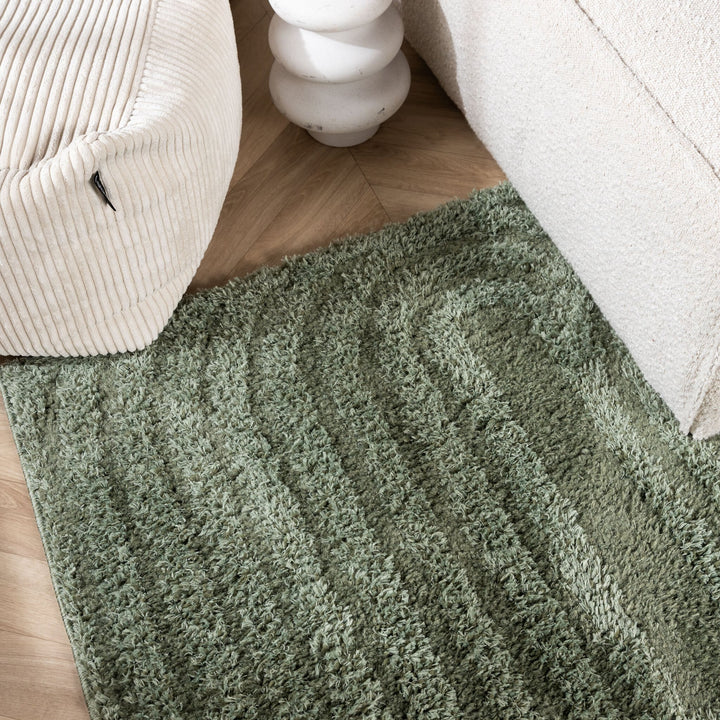 Corner of the green rug on a wood floor between a cream corduroy pouf and a sofa 3