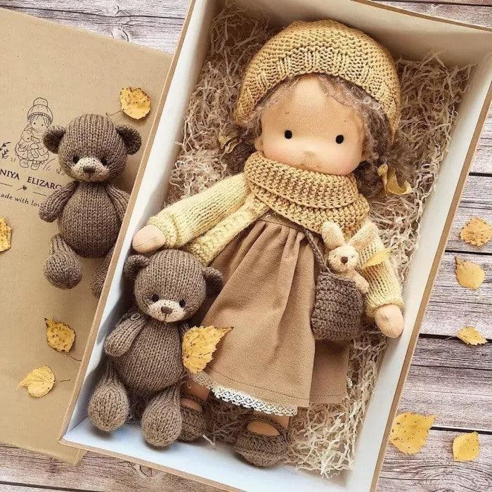 Doll in a mustard knit hat and scarf with brown dress, boxed beside two knitted teddy bears among autumn leaves 6