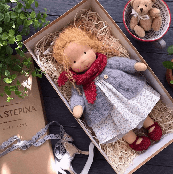 Doll with ginger curls, red scarf, gray cardigan and red shoes in a gift box surrounded by greenery 9
