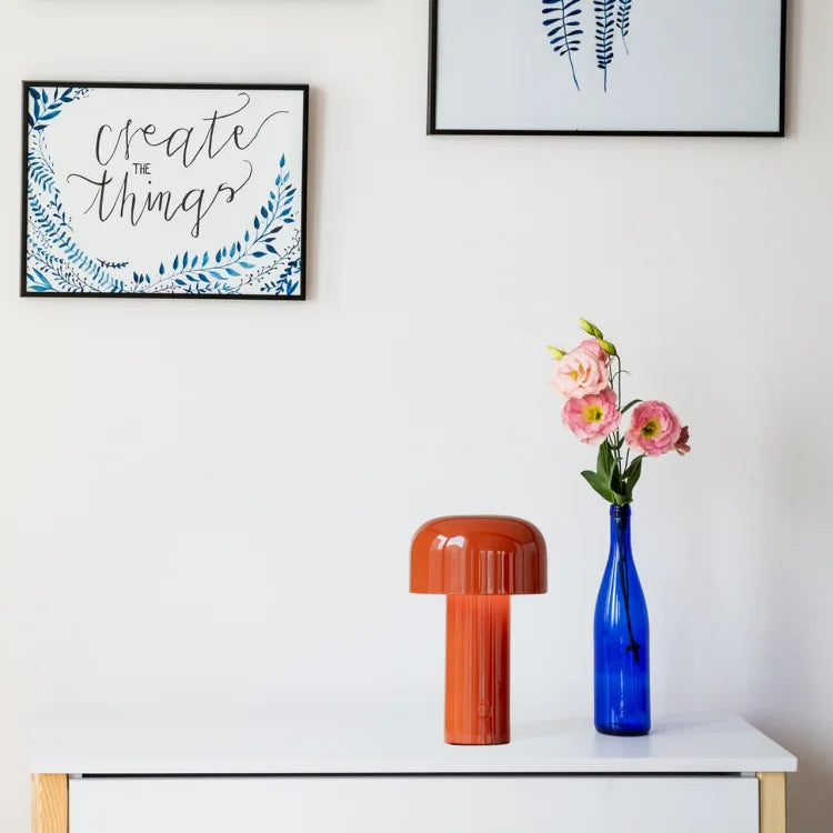 Red-orange lamp on a white dresser beside a blue bottle with pink flowers, with framed prints above 20